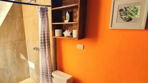 Un baño con pared naranja, un inodoro, una ducha con cortina, un lavabo y un estante con artículos de aseo.