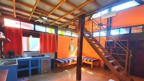 Una habitación pequeña y rústica con techo de madera, una escalera, una pequeña cocina y una zona de estar con bancos.