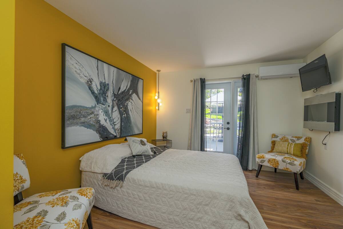 A bedroom with a bed, a chair, a painting, and an air conditioning unit.