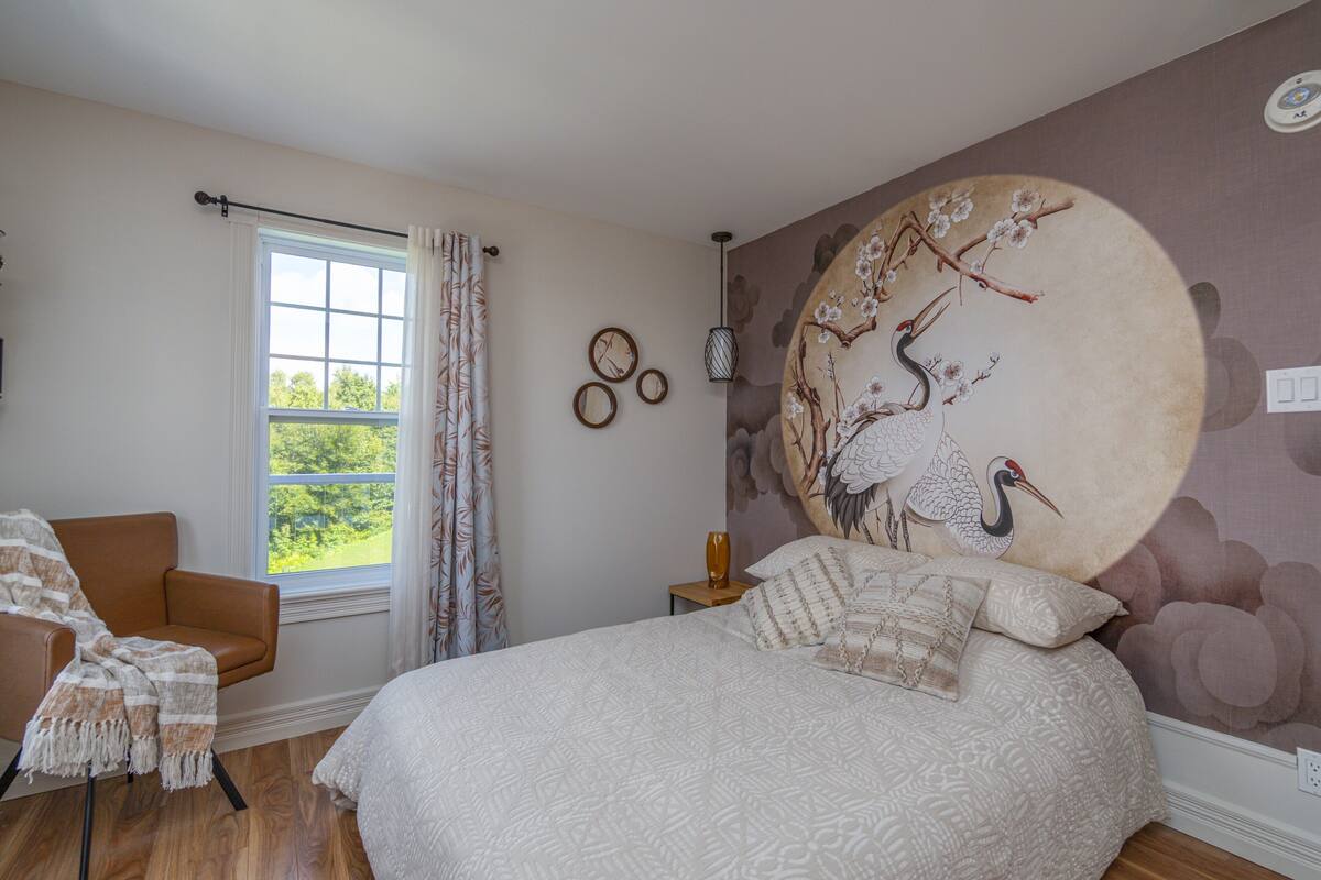 A bedroom with a large bed, a window with curtains, a chair with a blanket, and a wall with a mural.
