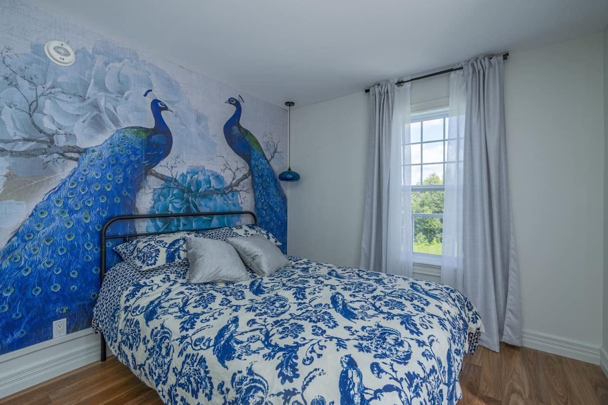 A bedroom with a bed, a peacock mural, a window with curtains, and a hanging light fixture.