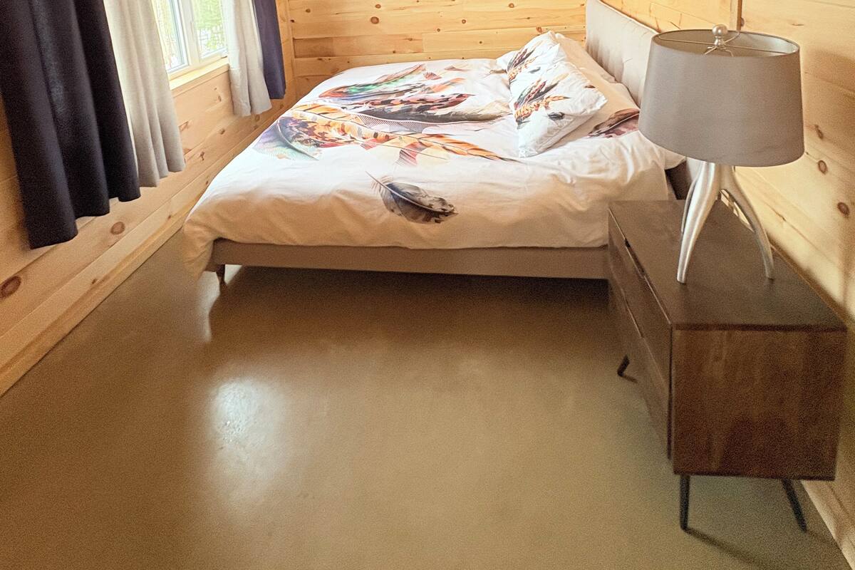 A wooden cabin room with a bed, a nightstand, and a window with curtains.