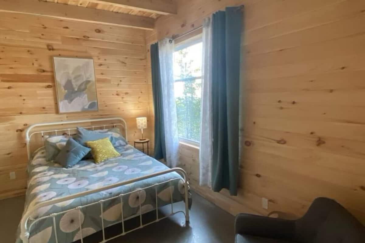 A wooden cabin room with a bed, a chair, a window with curtains, and a small table with a lamp.