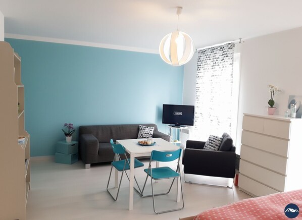Een compacte kamer met een blauwe wand, een witte tafel met stoelen, een grijze bank, een televisie en een garderobe.