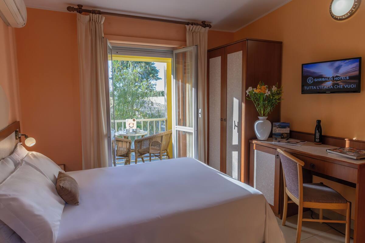Une chambre d’hôtel comprenant un lit, un bureau, une chaise, une télévision et un balcon équipé de fauteuils en osier.