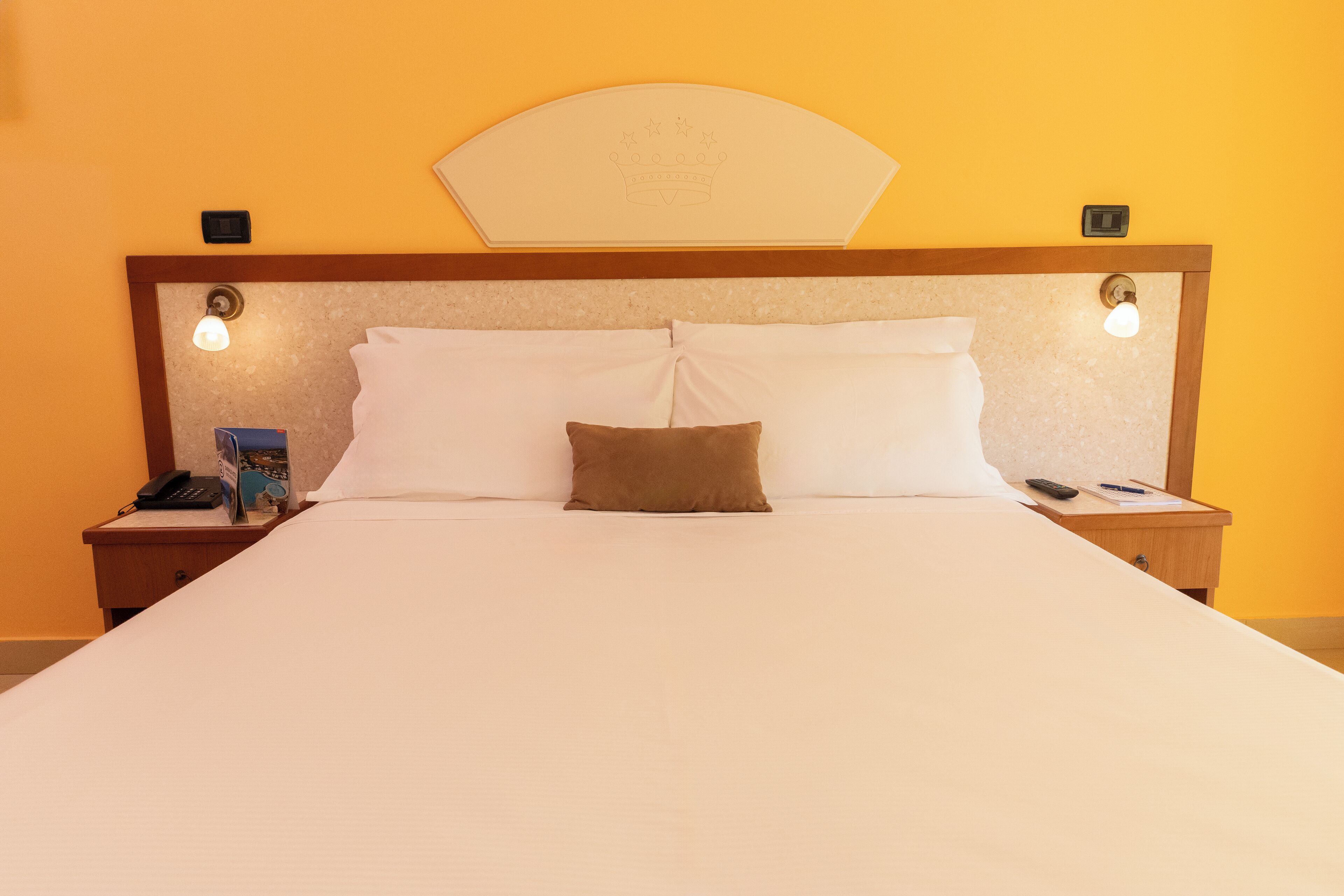 Une chambre d’hôtel avec un grand lit, deux tables de chevet, une petite table d’appoint, une tête de lit ornée d’un motif en forme de couronne et un luminaire fixé au mur.
