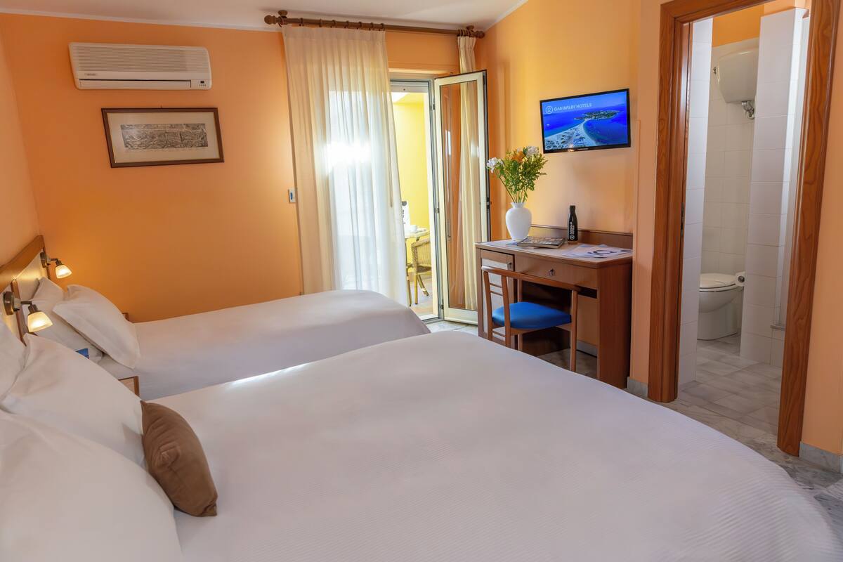 Une chambre d’hôtel avec deux lits, un bureau avec une chaise, une télévision et une salle de bain visible à travers une porte ouverte.