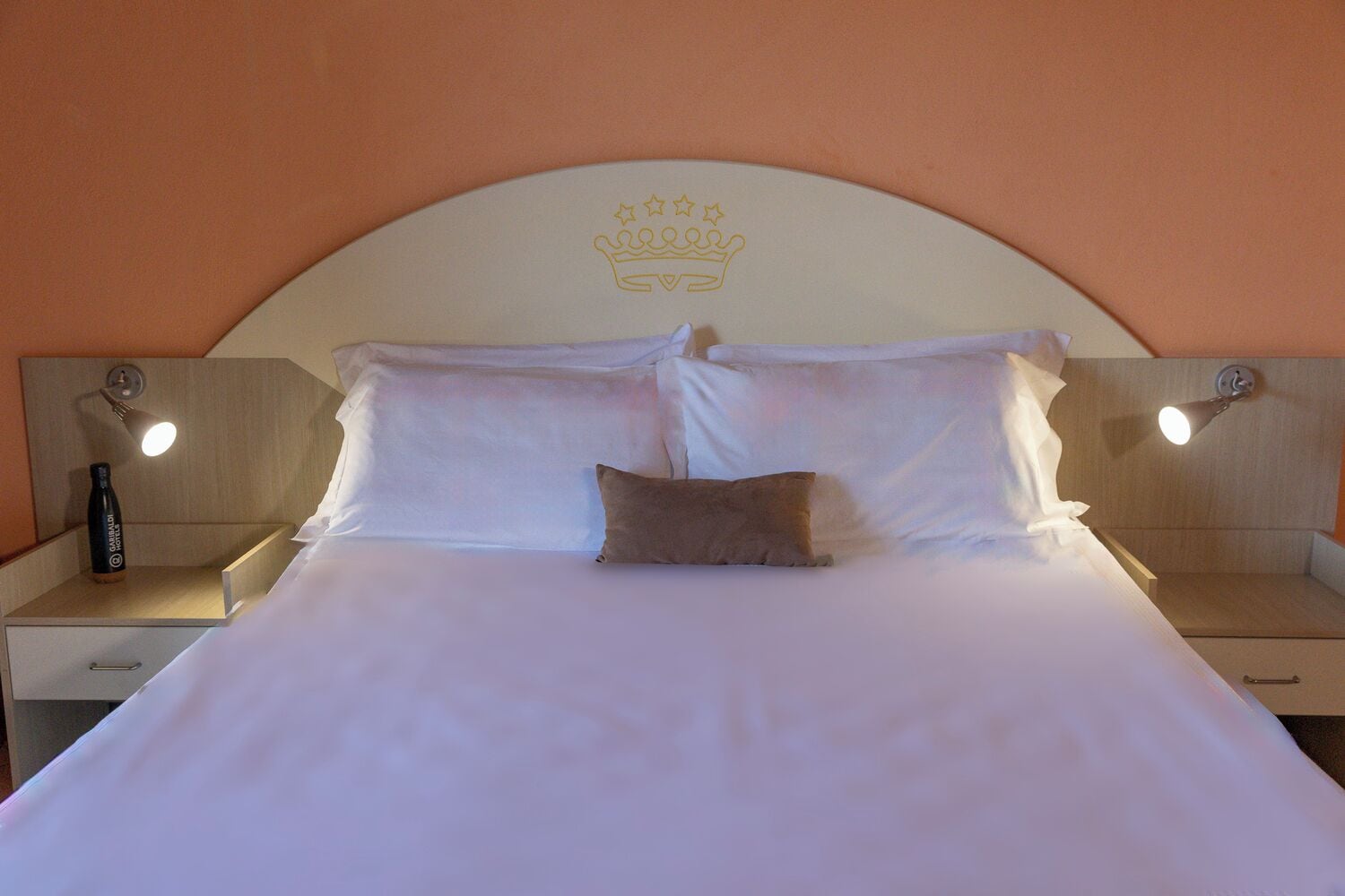 A hotel room with a large bed, two bedside tables, a bottle of water, and a headboard with a crown design.