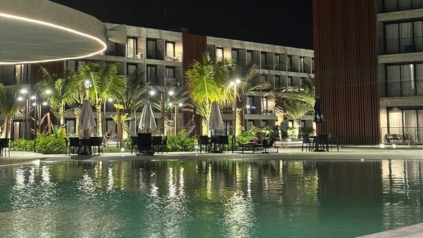 Vue nocturne d’un espace piscine d’hôtel, avec des chaises longues et des parasols, entouré d’immeubles dotés de balcons et de grandes fenêtres.