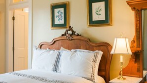 A neatly made bed with white linens and striped pillows, a wooden headboard, a bedside table with a lamp, and two framed botanical prints on the wall.