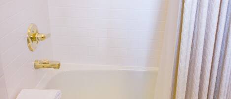 A white tiled bathroom with a bathtub, a gold faucet, and a stack of white towels.
