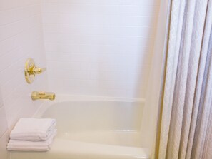 A white tiled bathroom with a bathtub, a gold faucet, and a stack of white towels. - Room 1-Double room-Private Bathroom-Standard (Fish Creek)