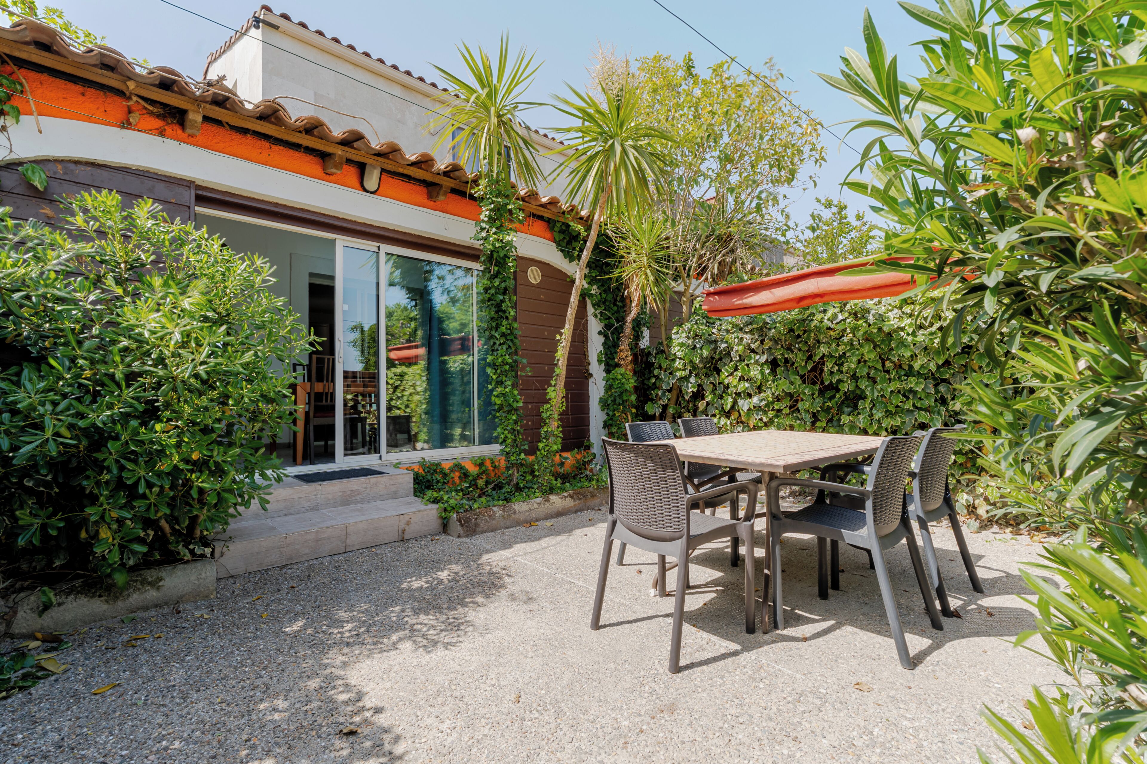 Une terrasse extérieure avec une table et des chaises, entourée de verdure et bordée d’un bâtiment dont le toit est recouvert de tuiles.