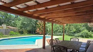 Eine überdachte Terrasse mit Pool, Holzdeck und Gartenmöbeln aus Metall.