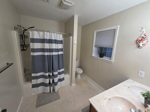 A bathroom with a shower curtain, toilet, sink, and a window with a plant. - Irene's Hideaway (Tisbury)
