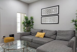 Ein Wohnzimmer mit einem grauen Sofa, einem Glastisch, einem gelben Kissen und zwei gerahmten abstrakten Kunstwerken an der Wand.