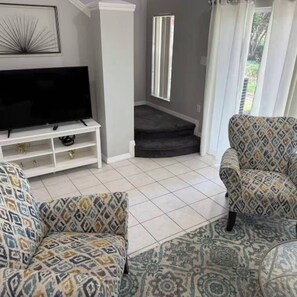 Ein Wohnzimmer mit gemusterten Sofas, einem Flachbildfernseher auf einem weißen Standfuß, einer Treppe und Blick ins Freie.