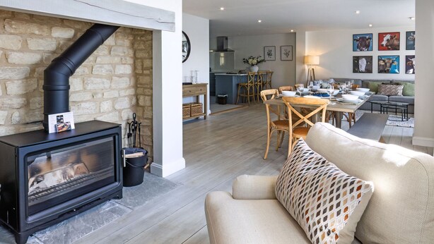 A cozy living room with a stone wall, a fireplace, a sofa, and a dining area with chairs and a table. - Mill Village 71/72, Milly Cottage - Lakeside, Onsite Facilities, Pool, Sleeps 11 (Somerford Keynes)