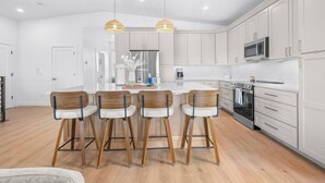 Une cuisine moderne avec des meubles blancs, un îlot central doté de tabourets de bar, des appareils en acier inoxydable et un éclairage suspendu.