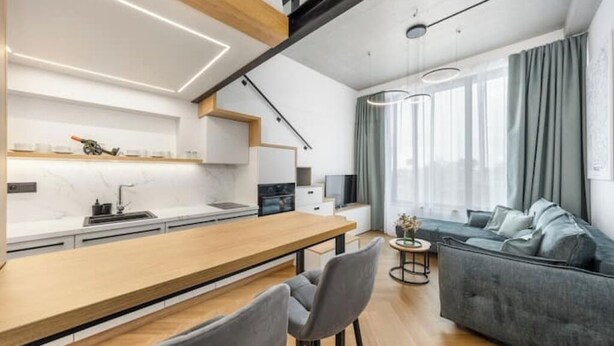 Une cuisine moderne avec un plan de travail en bois et des tabourets de bar, un canapé gris et une table à manger dans un espace de vie lumineux doté de grandes fenêtres.