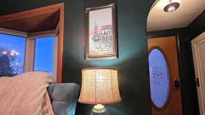 A room with a dark green wall, a framed picture, a table lamp, a wooden floor, and a window with a view of trees. - Charming Cozy Home - Relax & Recharge (Hartington)