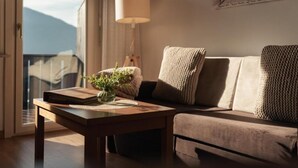 Een gezellige woonkamer met een bank, een bijzettafeltje met een boek en een plant, een lamp en een raam met uitzicht op bergen.