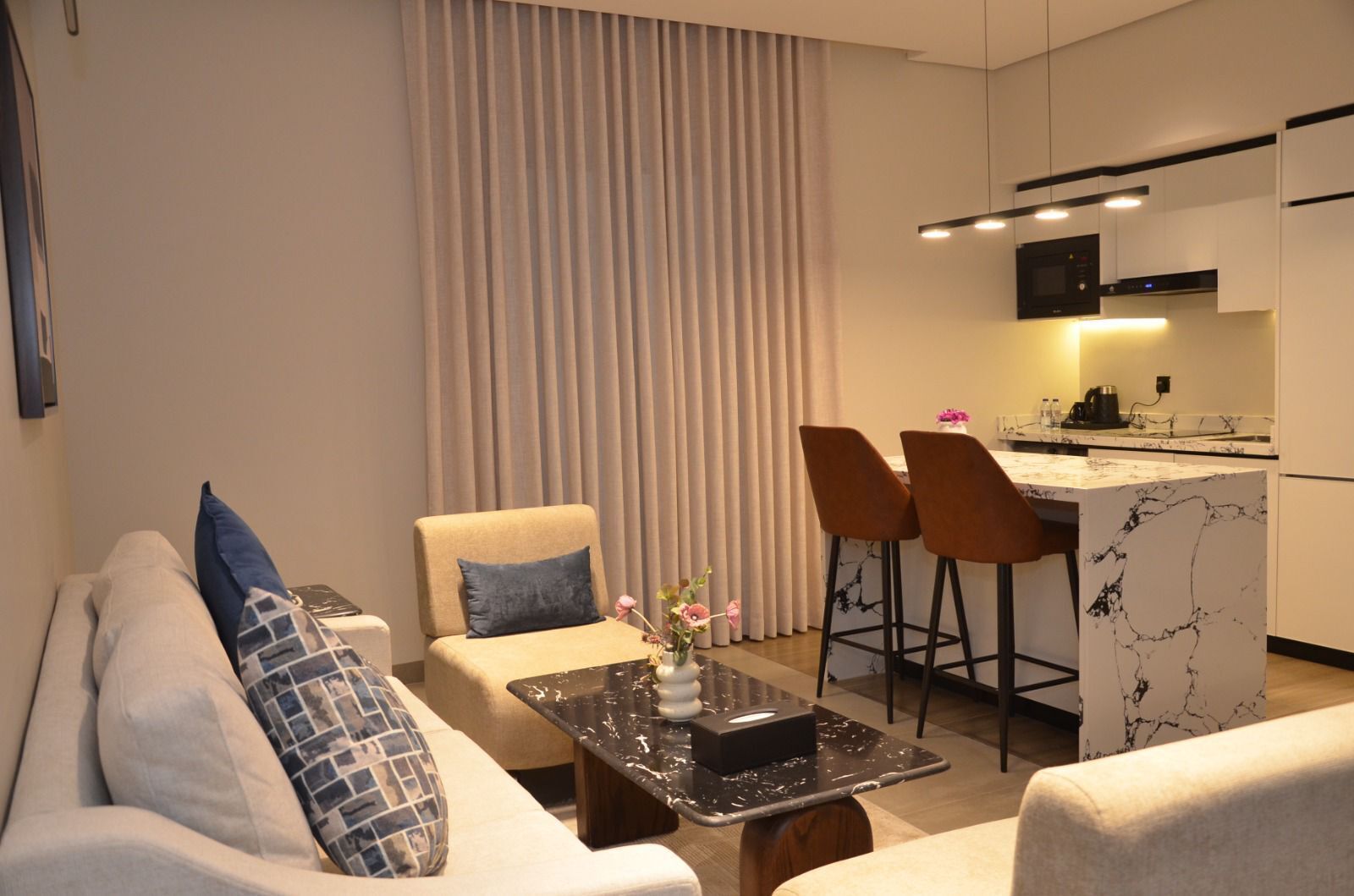 Un salon moderne comprenant un canapé, deux fauteuils, une table basse et un coin cuisine doté d’un comptoir en marbre et de tabourets de bar.
