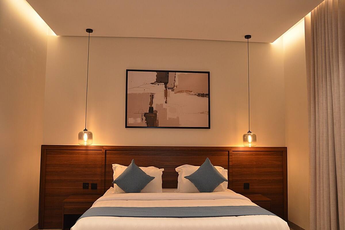 A hotel room with a large bed, two bedside tables, a wooden headboard, a framed abstract painting, and two pendant lights.
