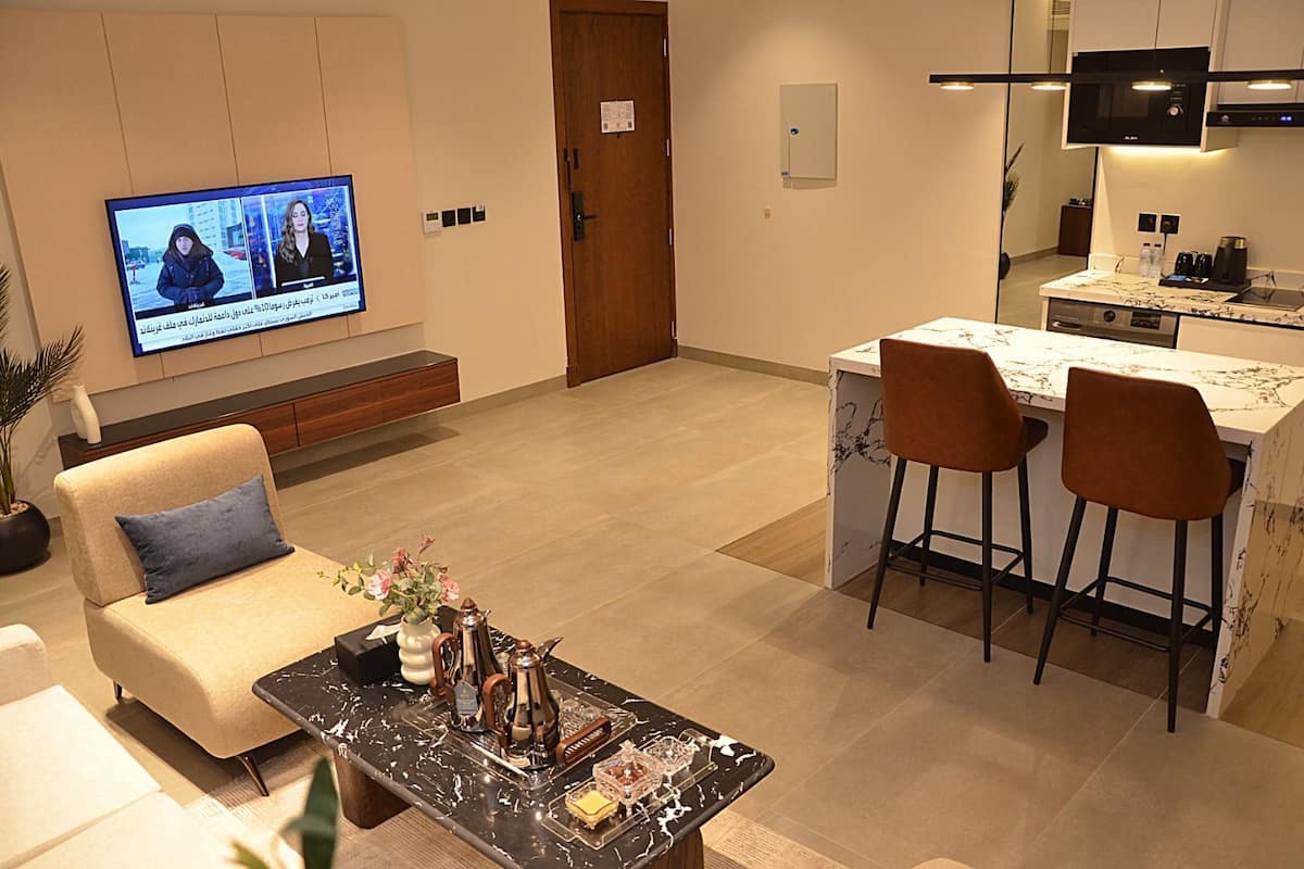 A modern living room with a sofa, a coffee table, and a TV displaying a news program. A kitchen area with a dining set is visible in the background.