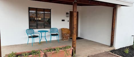A covered patio with two blue chairs and a table, a wooden chair, and terracotta planters with green plants.