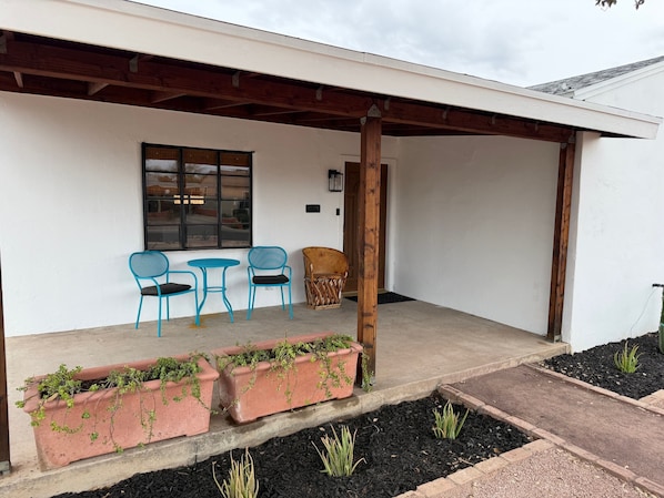 A covered patio with two blue chairs and a table, a wooden chair, and terracotta planters with green plants.