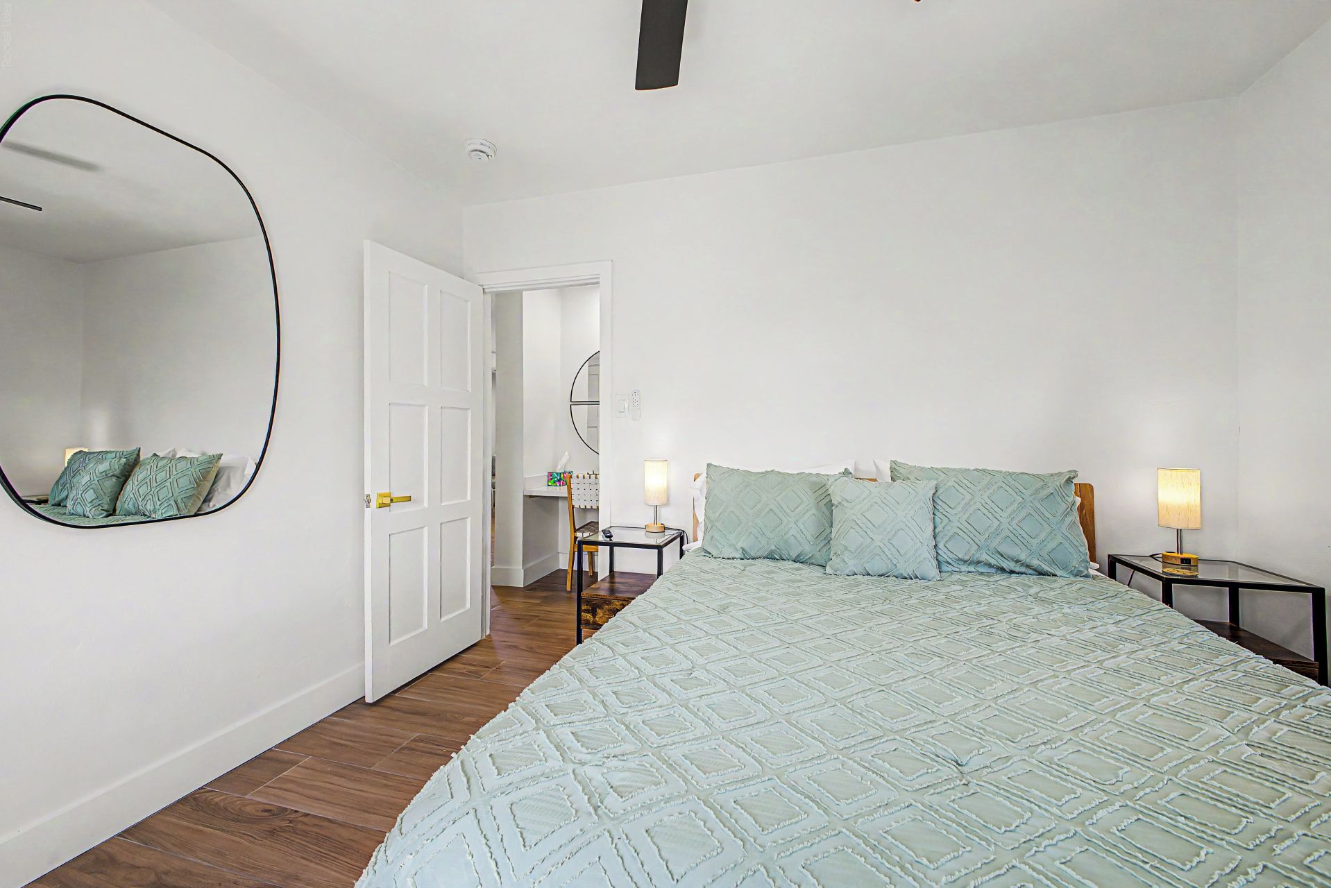 A bedroom with a bed, two bedside tables, a mirror, and a door leading to another room.