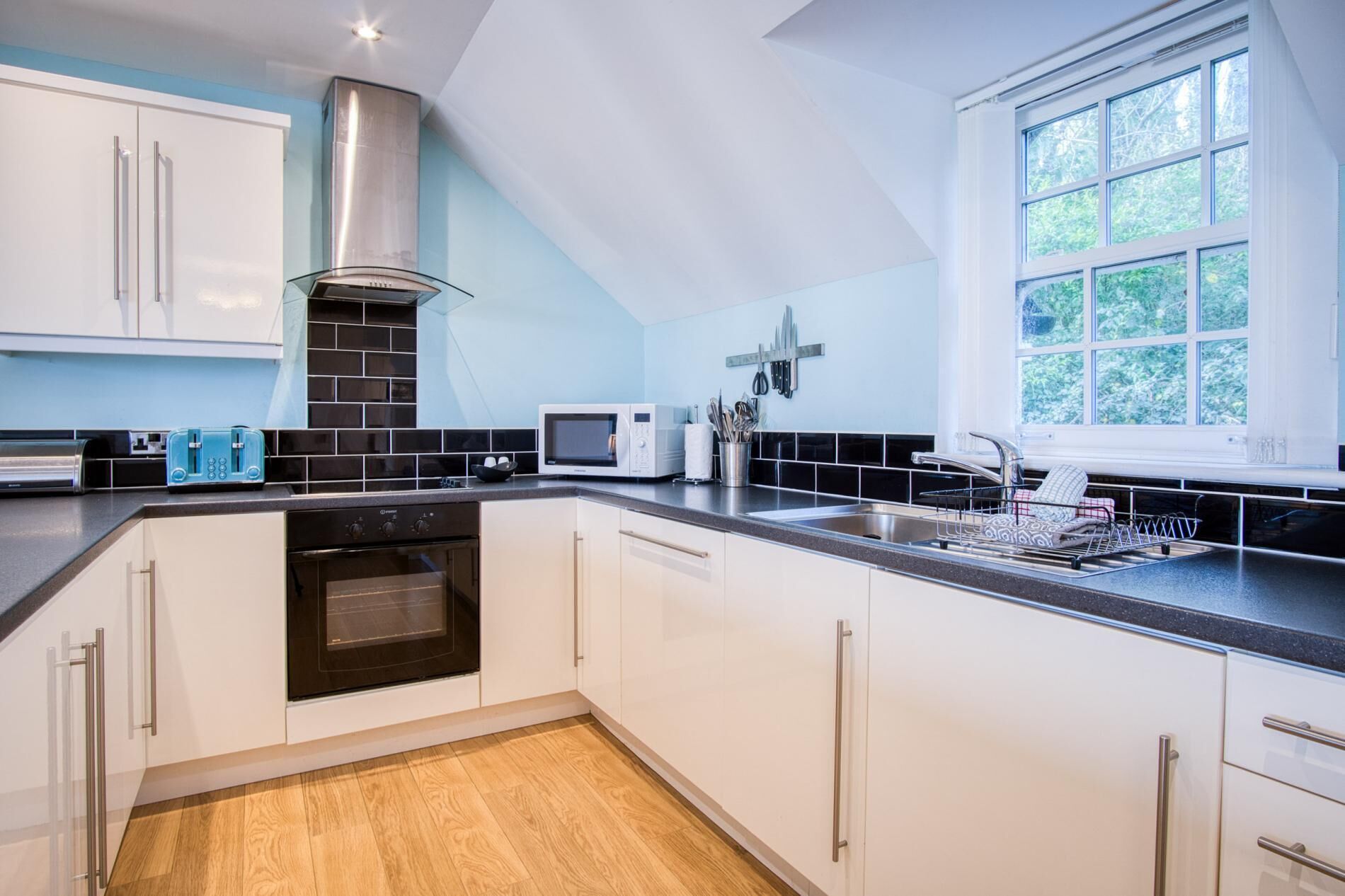 A modern kitchen with white cabinets, a stainless steel range hood, a microwave, and a window with a view of greenery.