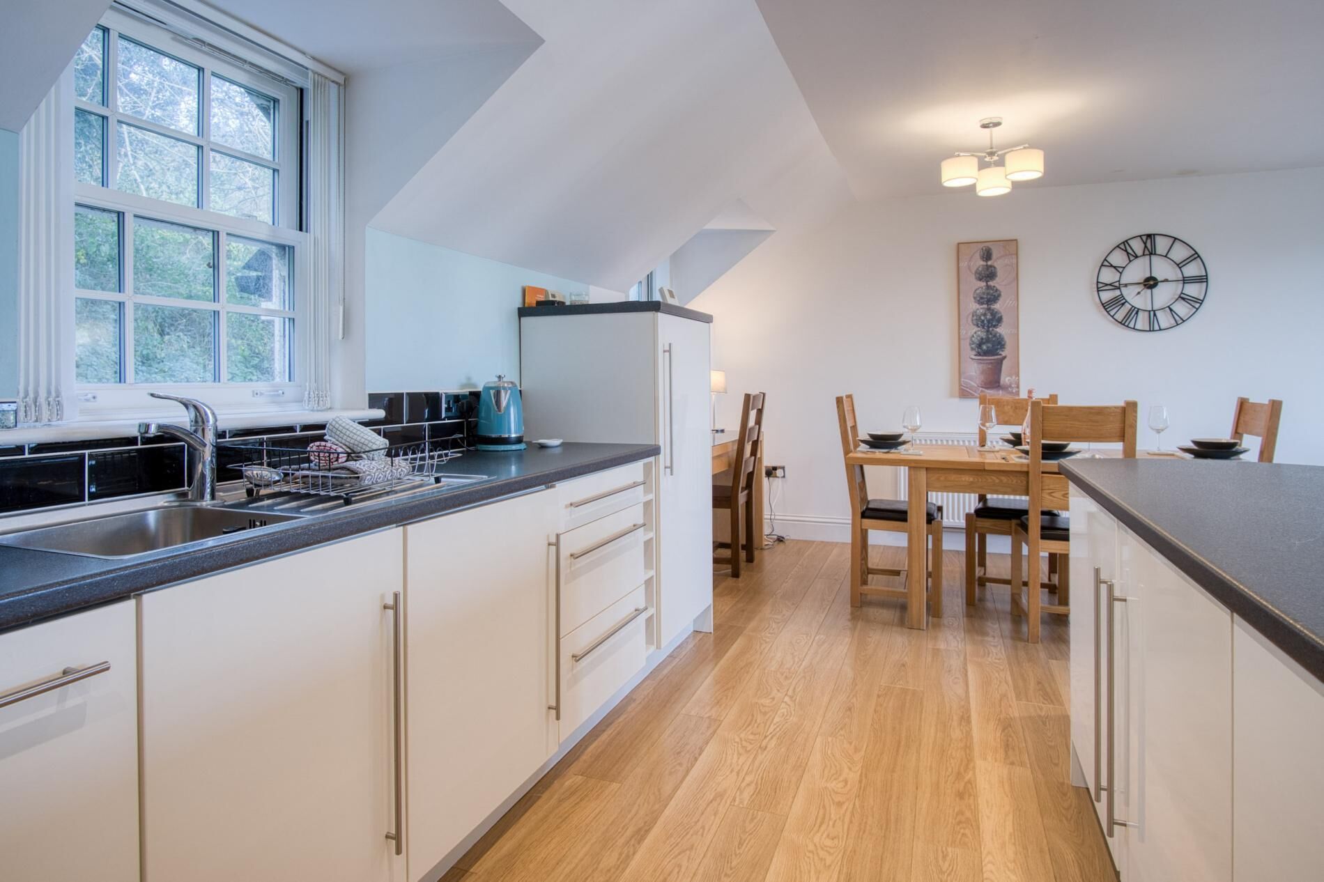 A modern kitchen with a large window, a sink, and a dining area with wooden chairs and a table.