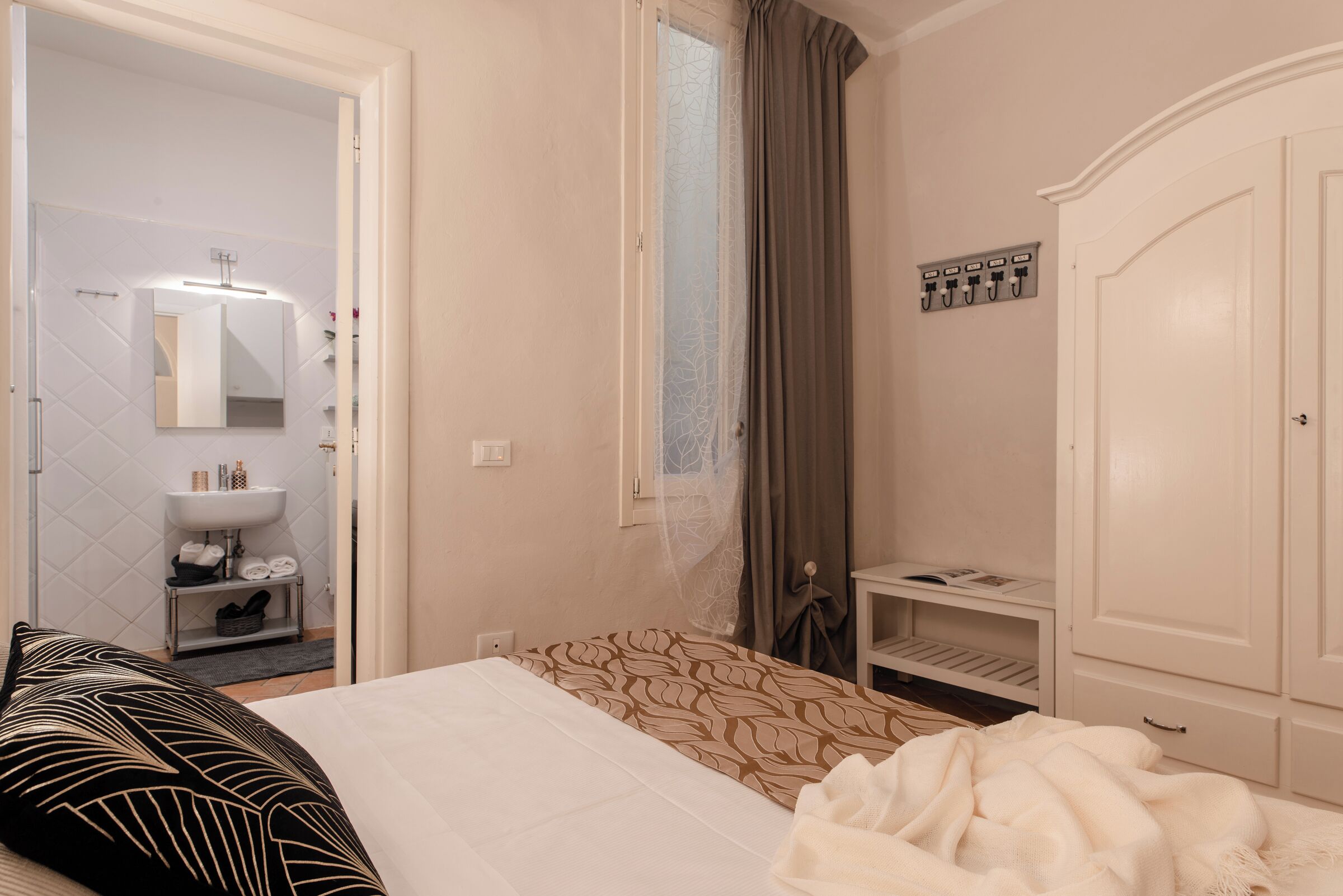Habitación de hotel con cama, mesita de noche, armario, baño con lavamanos y espejo, y ventana con cortinas.