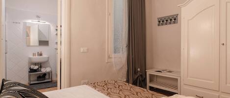 Habitación de hotel con cama, mesita de noche, armario, baño con lavamanos y espejo, y ventana con cortinas.