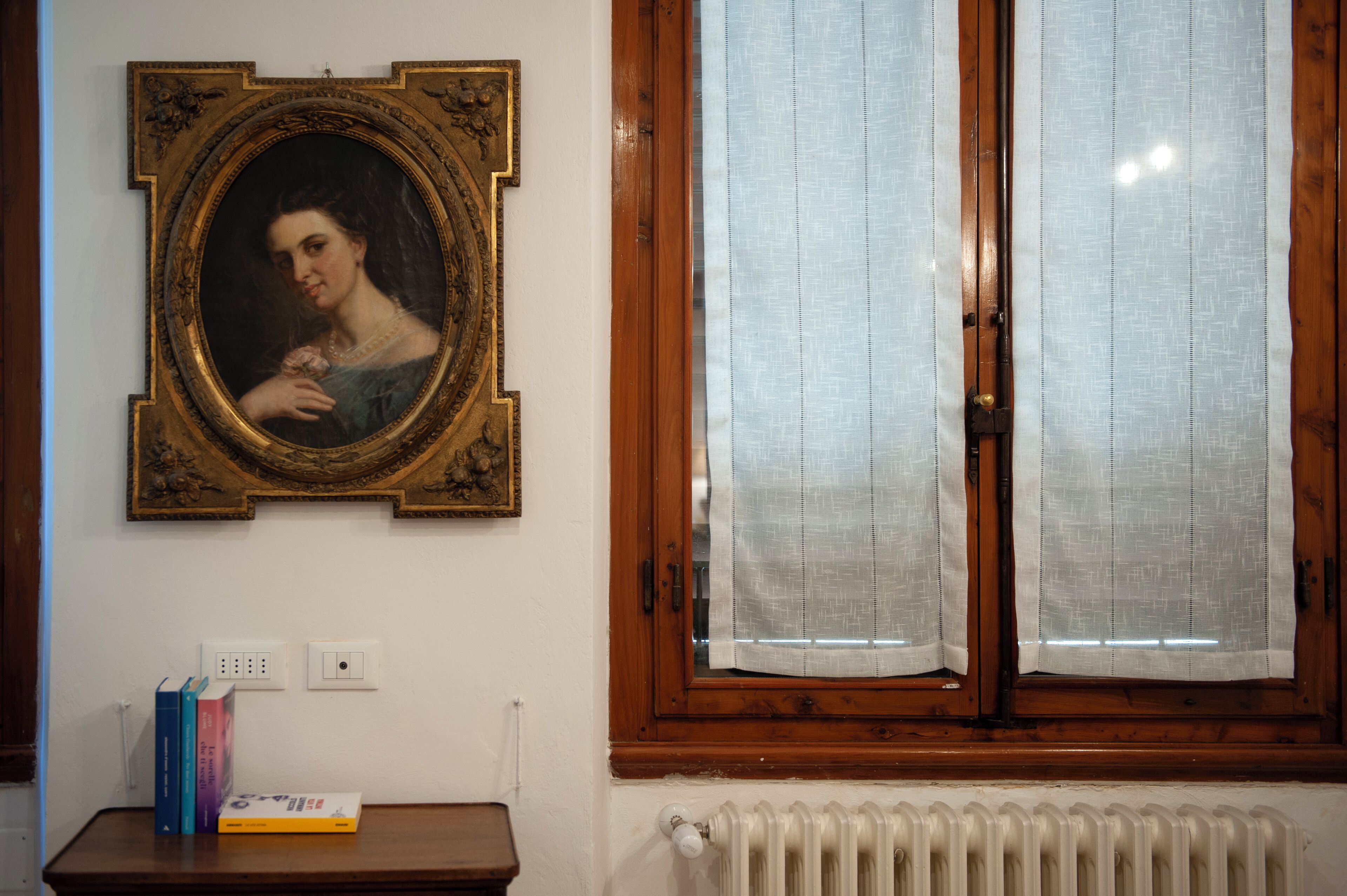 Una habitación con un retrato enmarcado en la pared, una ventana con cortinas, un radiador y una pequeña mesa con libros y una caja.