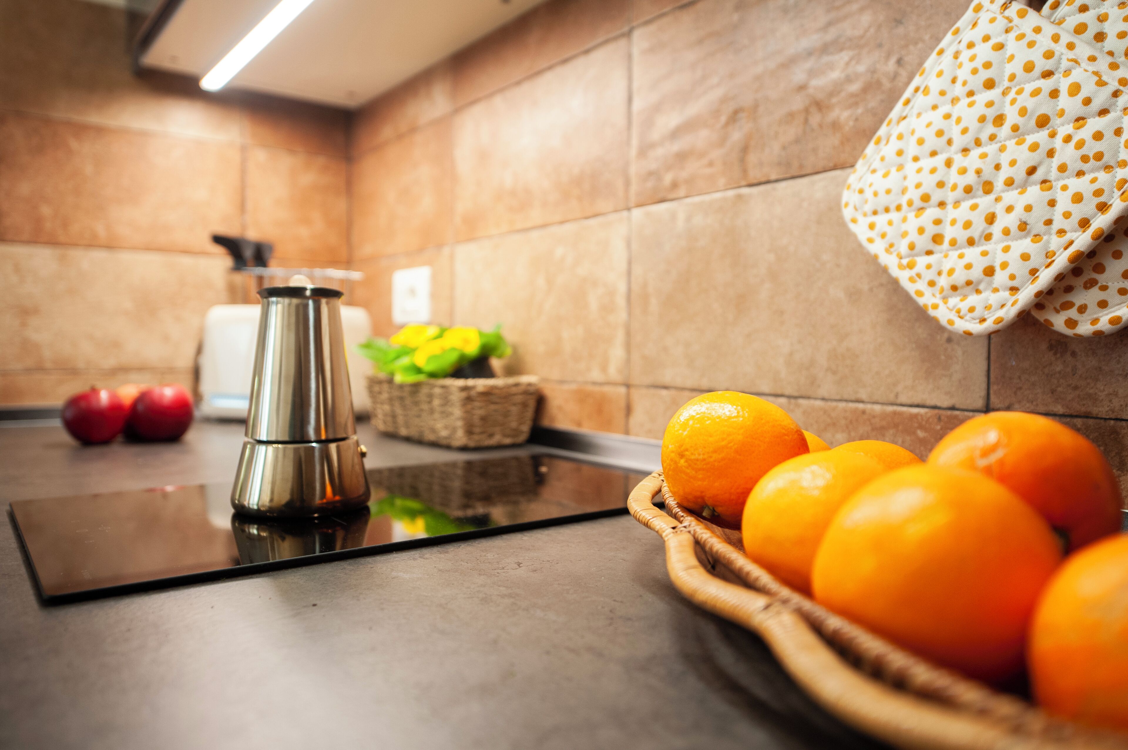 A kitchen with a stainless steel kettle, a basket of oranges, and a polka-dotted cloth hanging on the wall.