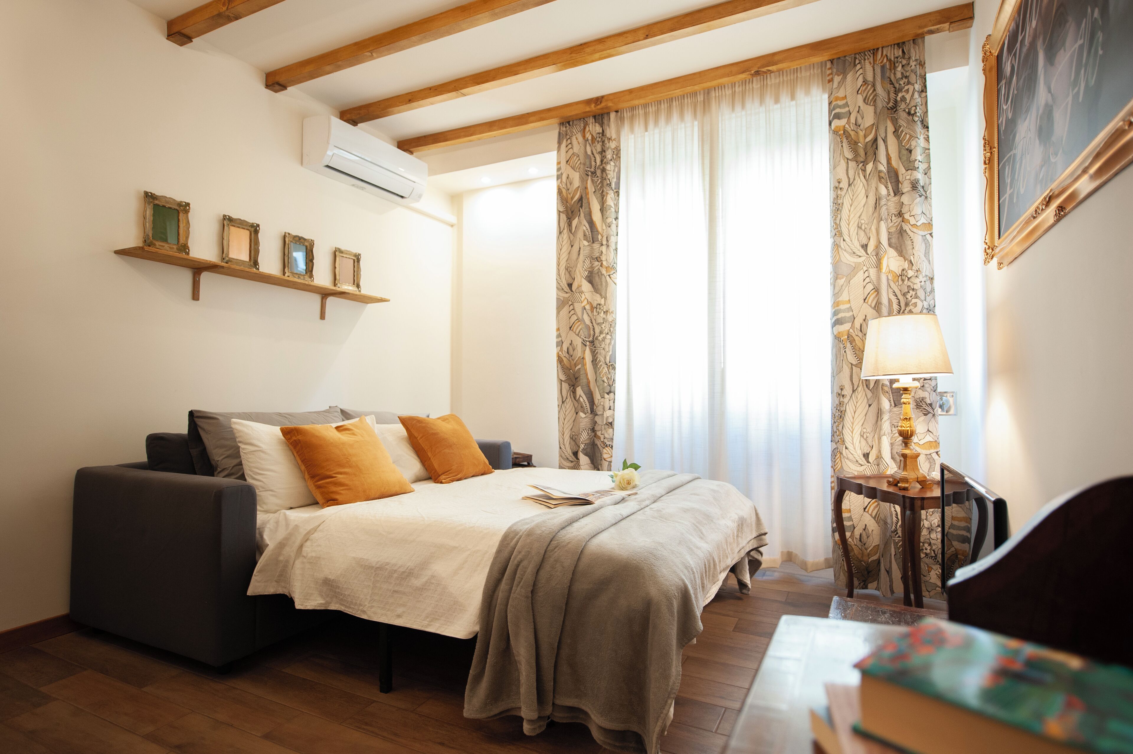 A bedroom with a bed, a sofa, a table with a lamp, and a wall-mounted air conditioner.