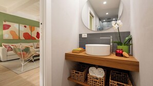 Un baño con un espejo redondo, un lavabo y una orquídea en maceta. Se ve un perchero con toallas dobladas y una pequeña planta en el mostrador.