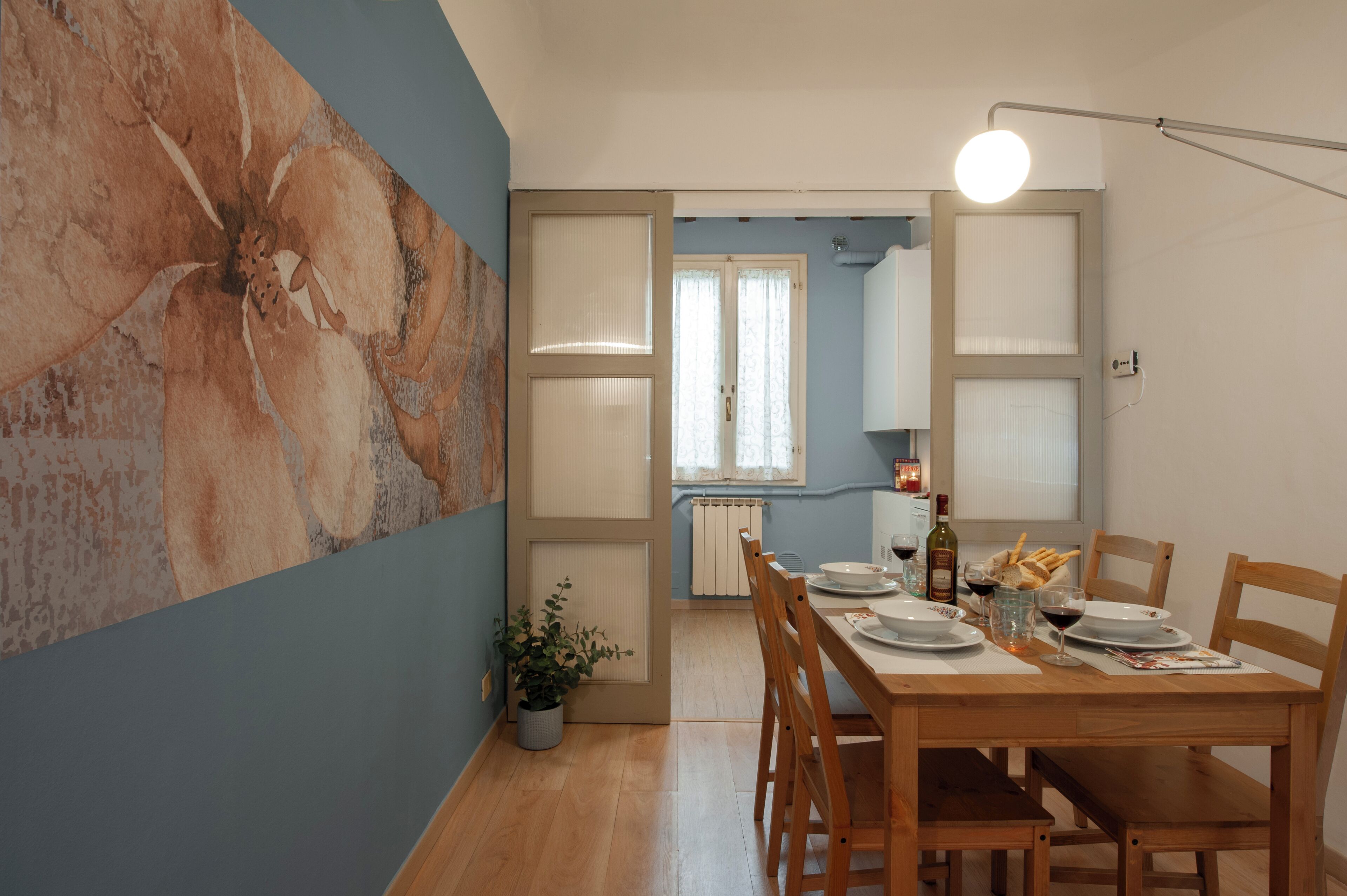 A dining area with a wooden table set for two, a potted plant, and a bottle of wine.