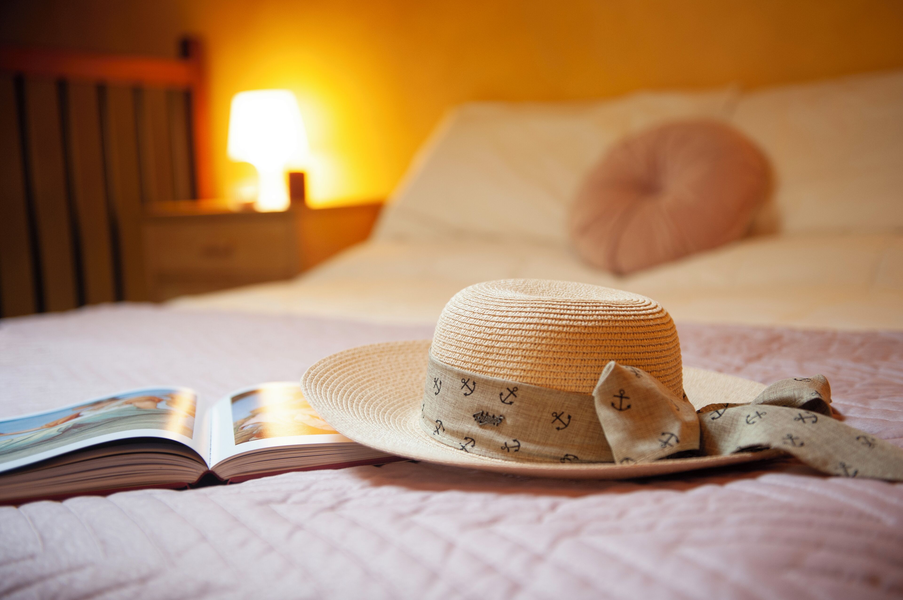 Una cama con un sombrero y un libro, una lámpara de noche y un almohadón.