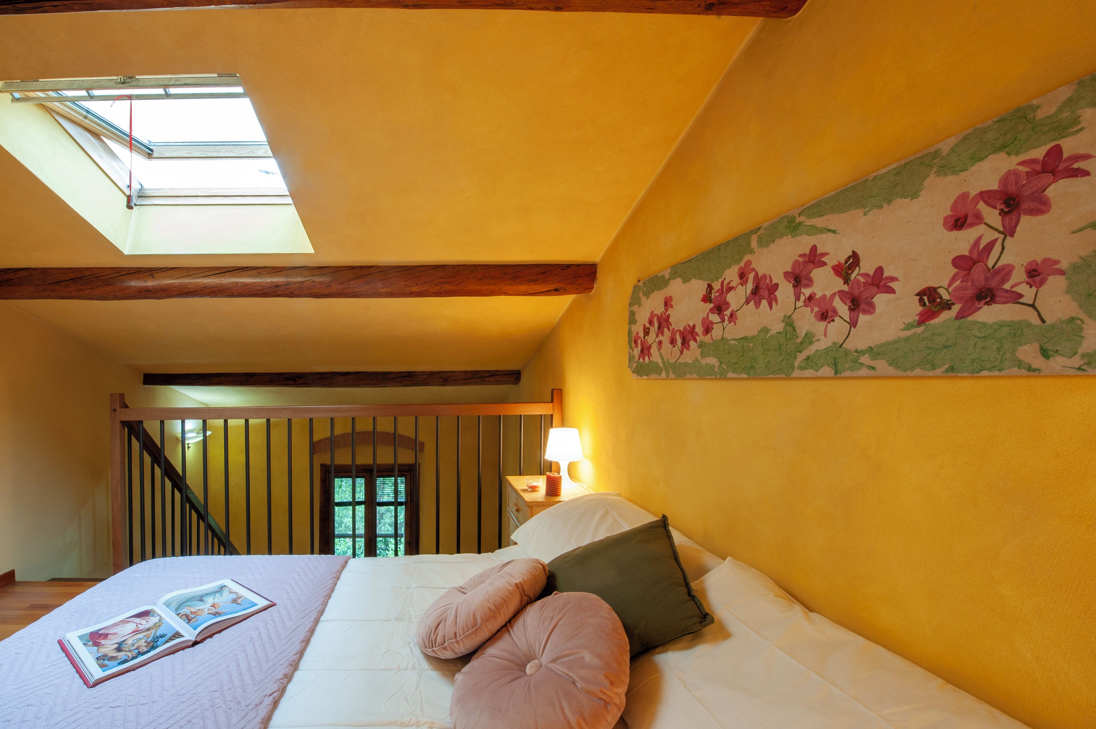 Una camera da letto con finestra a lucernario, un letto con cuscini, un comodino con una lampada e una decorazione floreale a parete.