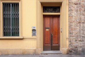 A building with a yellow facade, a stone wall, and a wooden double door with a metal handle. - Cate Apartment - Flo Apartments (Florence)