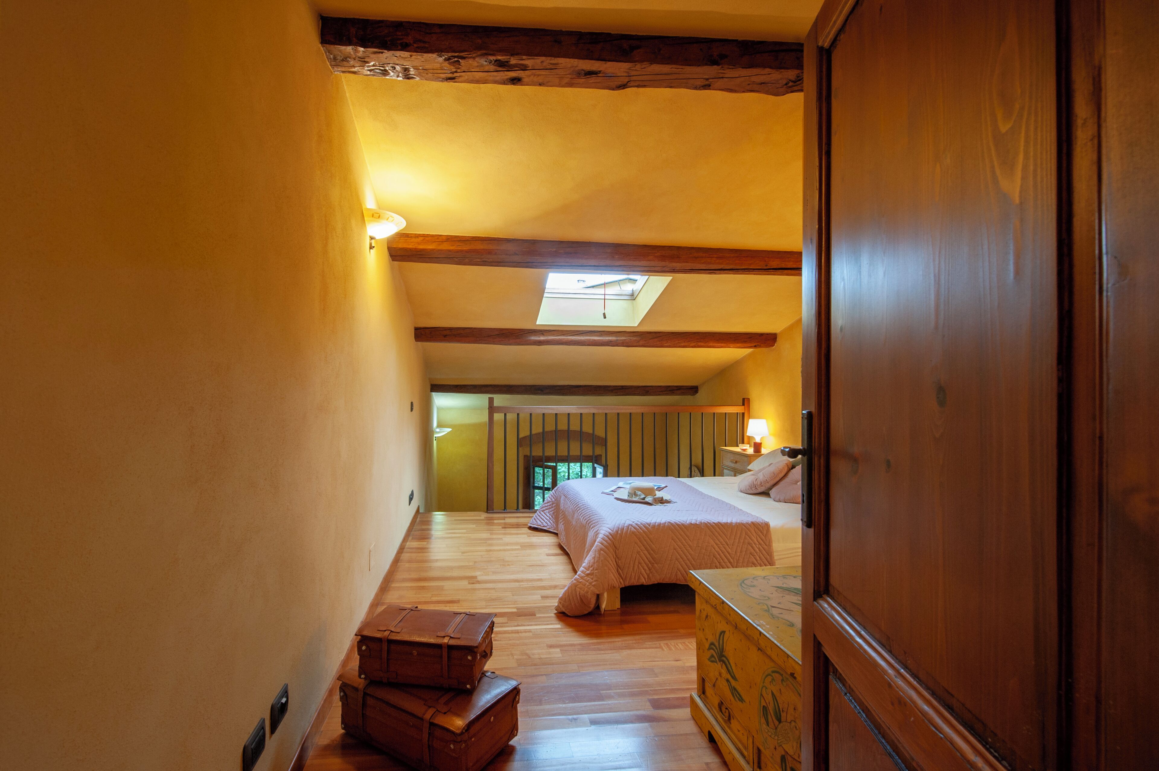 Un couloir étroit mène à une chambre dotée d’une lucarne, de poutres en bois et d’un lit recouvert d’une couverture blanche.