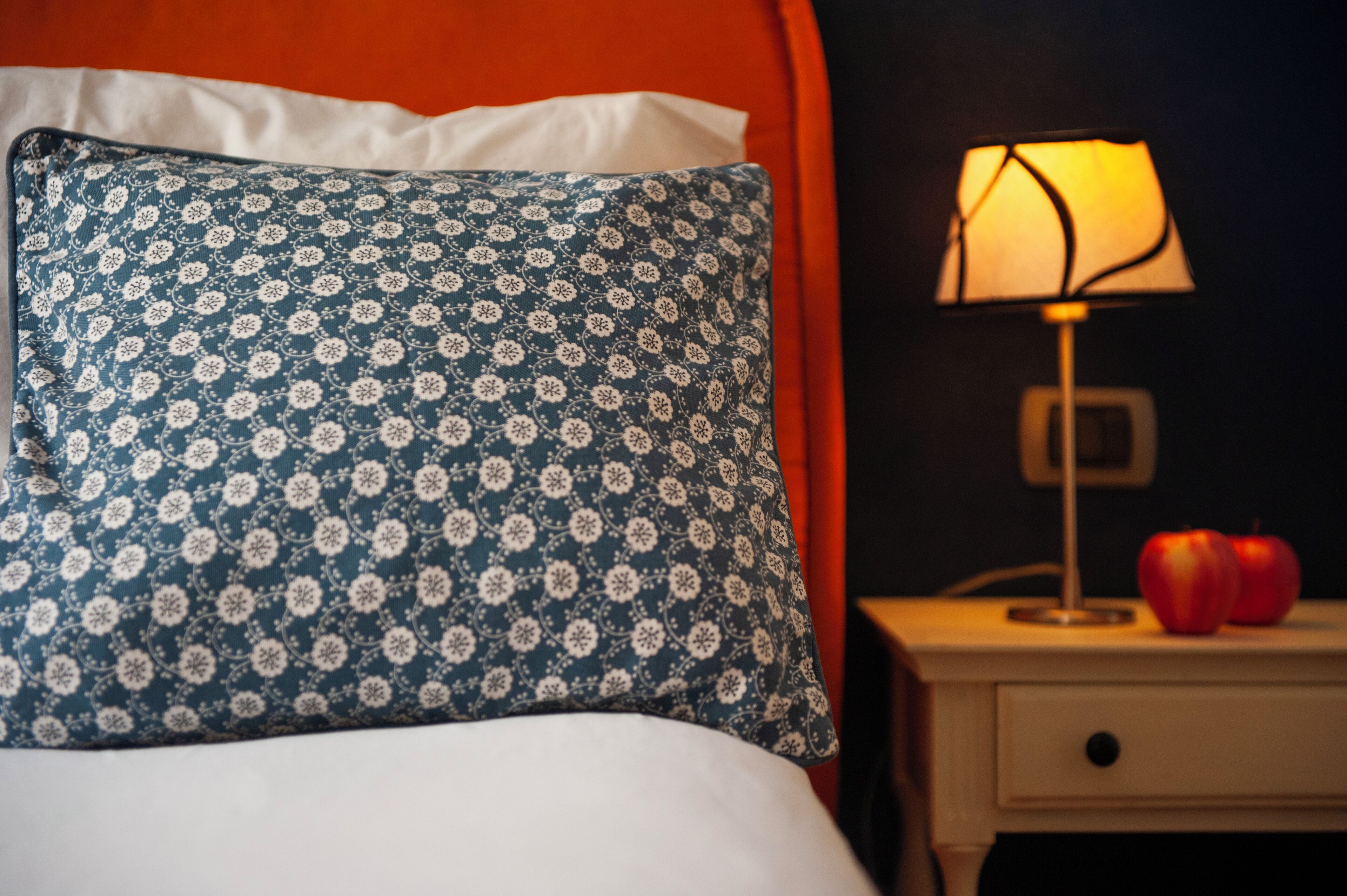 A bed with a patterned pillow, a bedside lamp, and two apples on a nightstand.