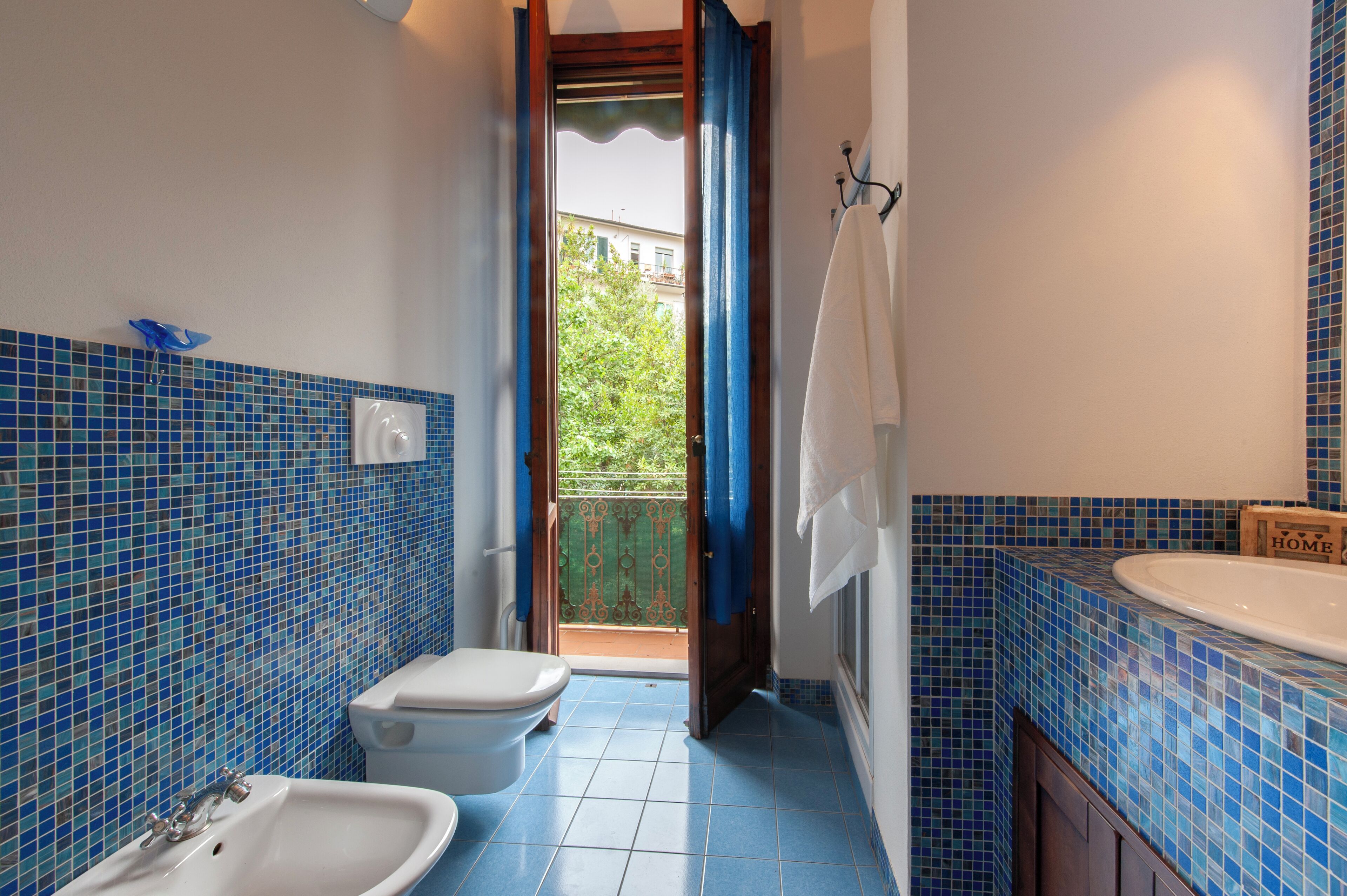 Ein Badezimmer mit blauen Mosaikfliesen, einem weißen Waschbecken, einer Toilette und einer Duschkabine mit Blick auf einen Balkon.