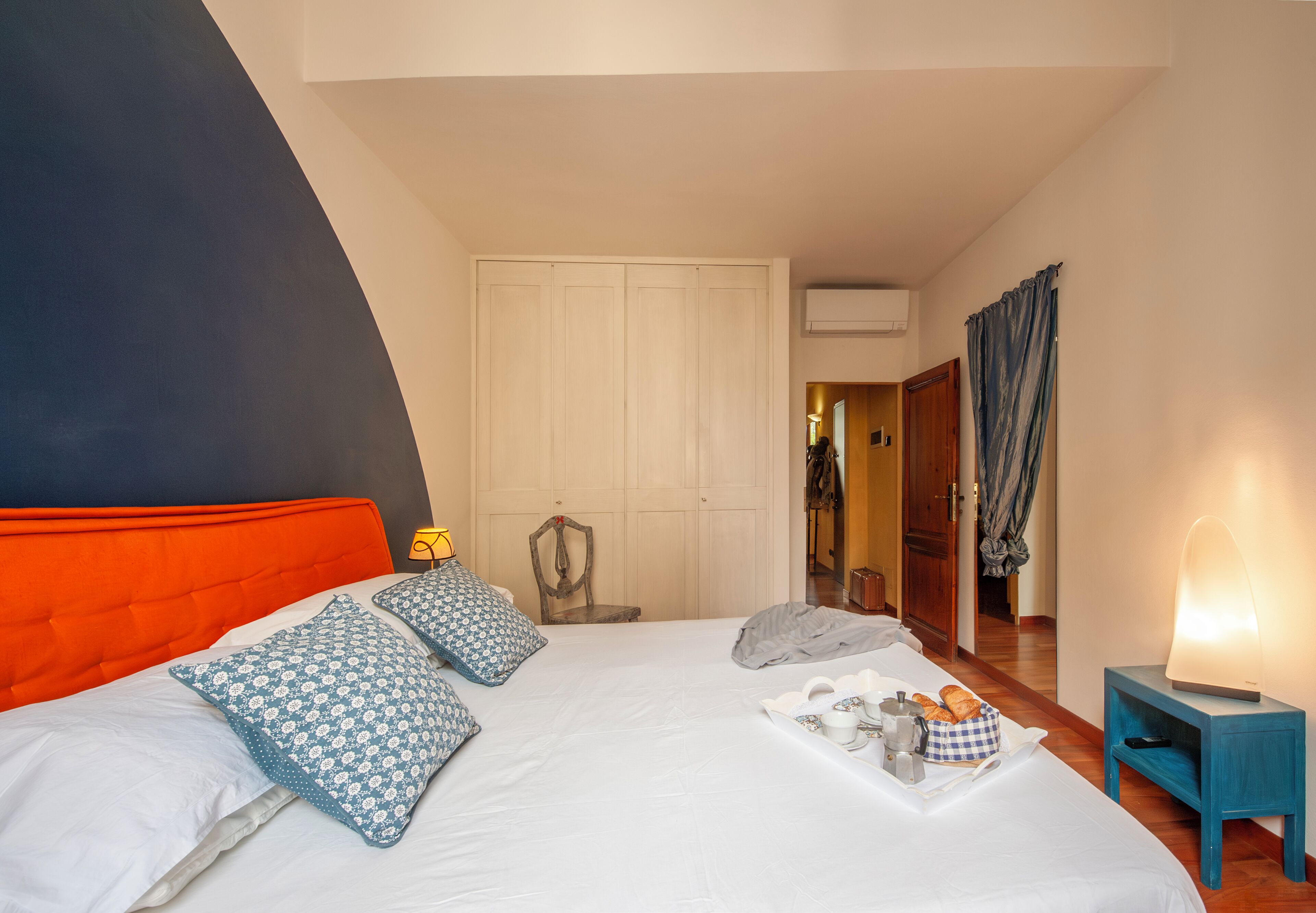 Un dormitorio con cama, almohadas, mesita de noche con lámpara, bandeja con alimentos para el desayuno y armario.