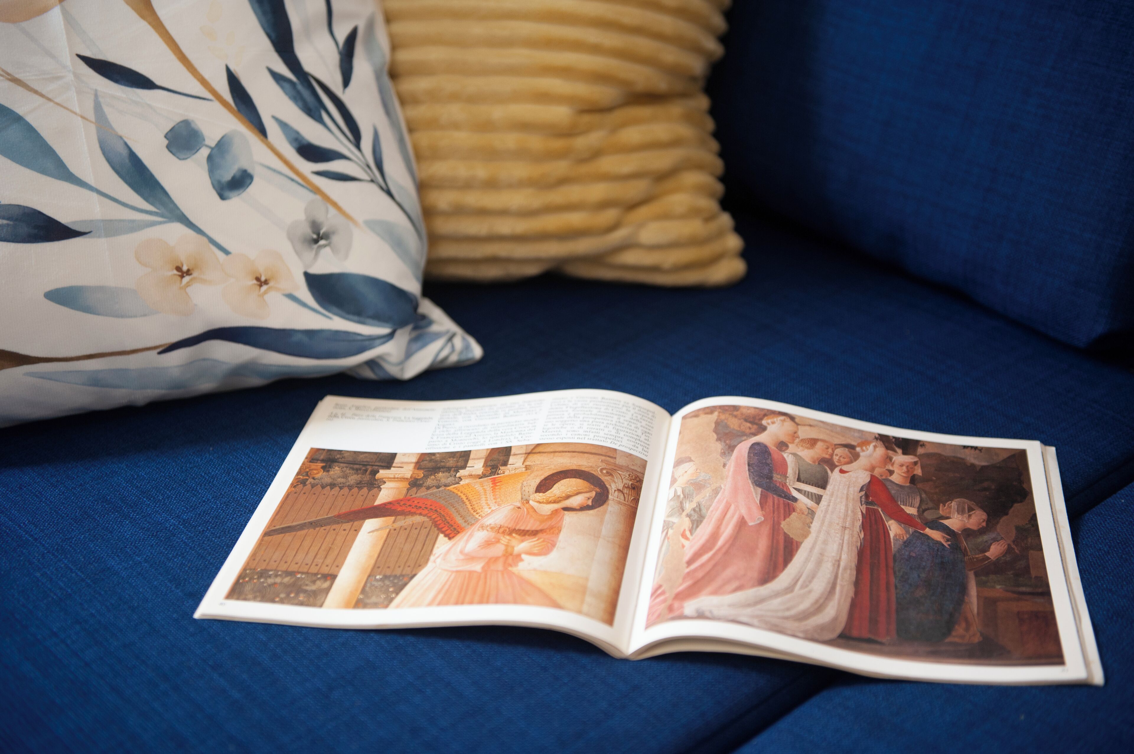 An open book with a page featuring a historical painting on a blue sofa with a floral pillow.
