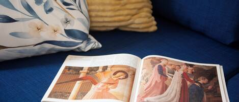 Un libro abierto con una página que muestra una pintura histórica sobre un sofá azul con un cojín floral.